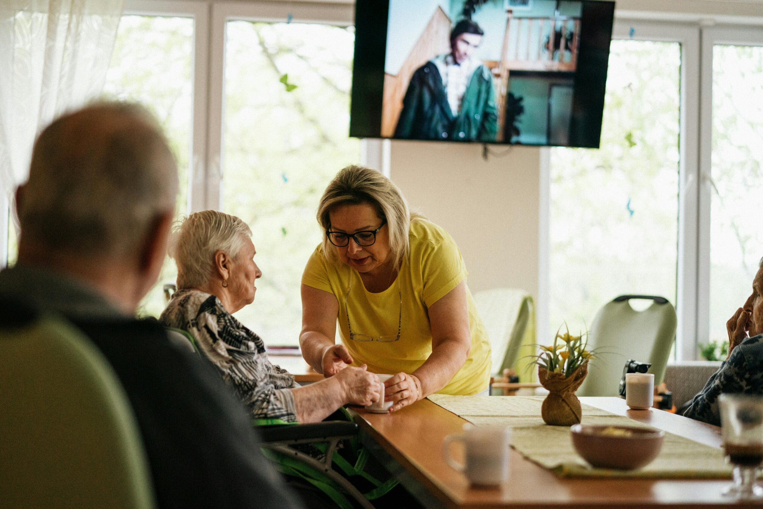 Caregiver assisting an elderly woman in a senior living facility with a TV playing in the background, showcasing commercial satellite TV for healthcare centers in the New York Metro Area.